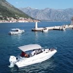 Drone view of a Good Hope Tours speed boat near Our Lady of the Rocks island, with Perast and mountains in the background and the sea surrounding them - Perast, Kotor, Blue Cave