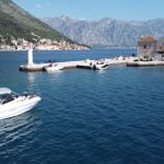 Drone view of a Good Hope Tours speed boat near Our Lady of the Rocks island, with Perast and mountains in the background and the sea surrounding them.